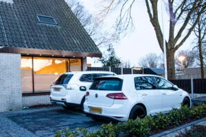 Oprit met donkere sierbestrating, twee witte auto's voor de carport en bakstenen woning