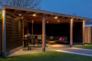 Donker houten buitenverblijf met spotjes en lampjes, BBQ, hoge tafel en loungeset, in de avondschemering