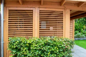Detail van een houten tuinberging met horizontale latten en luiken, achter een groene haag