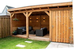 Houten overkapping met zwarte loungeset, hangende plant en wandlampen, aan de tuinrand
