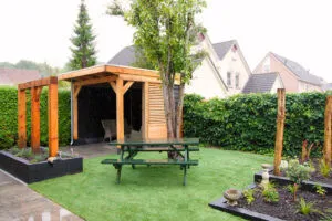 Tuin met houten overkapping, picknickbank op kunstgras, beukenhaag en plantvakken langs de rand