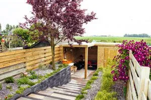 Tuin met houten overkapping, bloeiende sierboom, clematis tegen de schutting en uitzicht over de polder