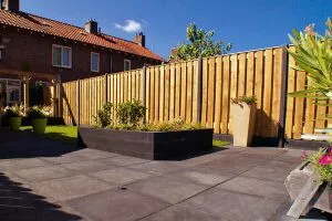 Voortuin met donkere bestrating, houten schutting en zwart stalen plantvak met beplanting