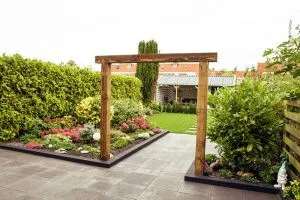 Houten pergolapoort als doorgang tussen twee plantvakken met bloeiende beplanting, taxushaag op de achtergrond